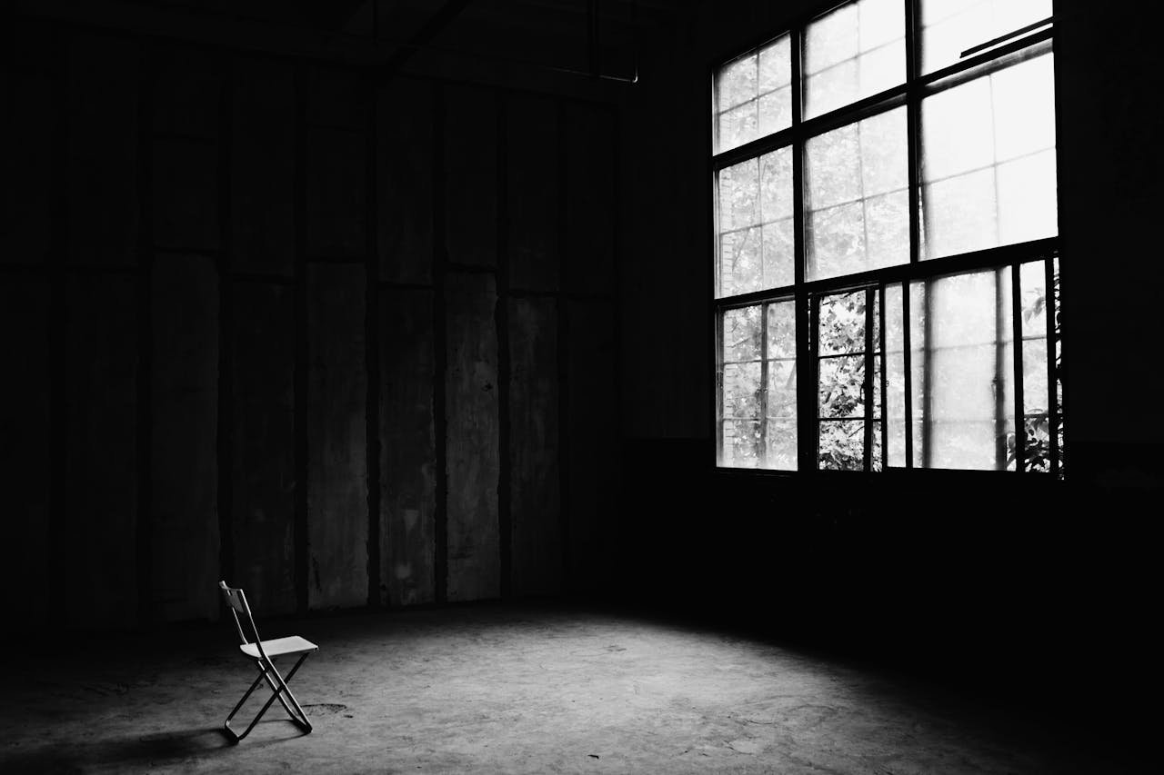 A dramatic black and white photo of an empty room with a single chair and large windows casting shadows.
